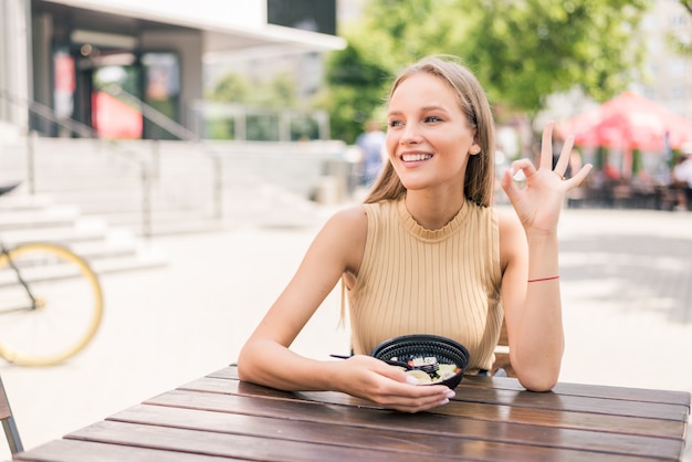 Молодая красивая женщина с хорошо жестом ест салат в кафе на открытом воздухе