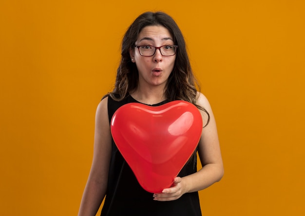 Giovane bella donna con palloncino rosso che sembra sorpresa festeggiando il giorno di san valentino sul muro arancione orange