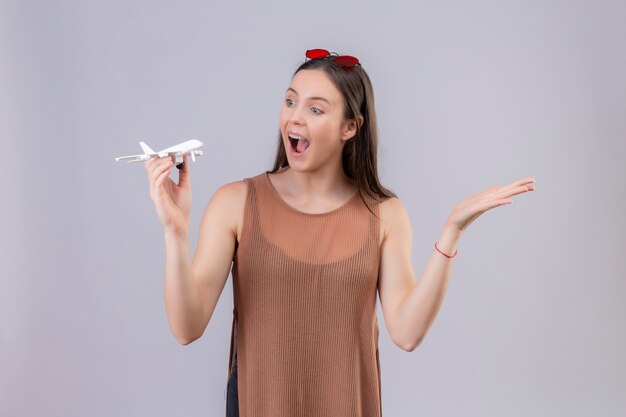 Young beautiful woman with red sunglasses on head holding toy airplane looking playful and happy with arm raised over white wall