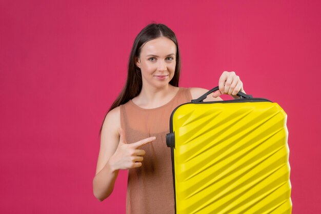 핑크 backgrpound 위에 서있는 카메라를보고 얼굴에 미소로 긍정적이고 행복을 손가락으로 가리키는 여행 가방을 가진 젊은 아름 다운 여자