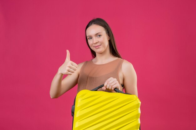 Young beautiful woman with travel suitcase with happy face smiling showing thumbs up over pink wall