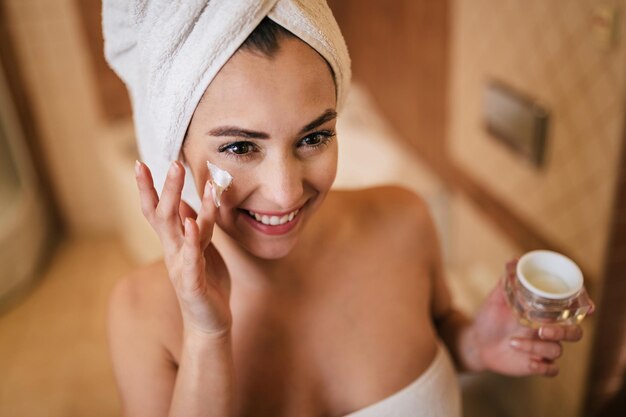 タオルに包まれた若い美しい女性がバスルームで彼女の顔に保湿剤を適用します