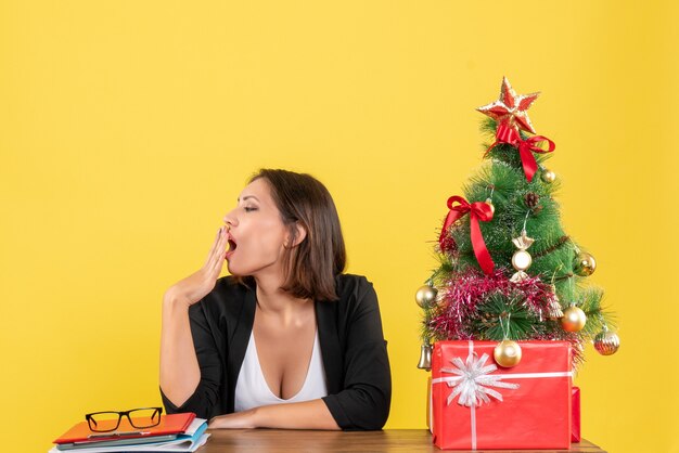 Young beautiful woman yawning sitting at a table near decorated Christmas tree at office on yellow 