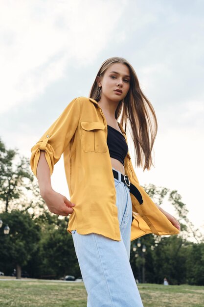 Free photo young beautiful woman in yellow shirt and jeans dreamily looking in camera while standing on lawn in city park