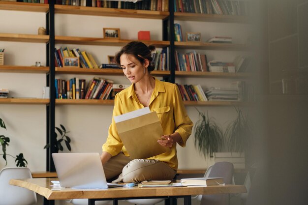Молодая красивая женщина в желтой рубашке сидит на столе с ноутбуком и бумагами, мечтательно работая в современном офисе