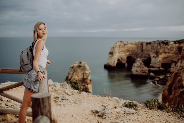 Молодая блондинка красоты Путешественник женщины смотря море и утес на красивом пляже Португалии.