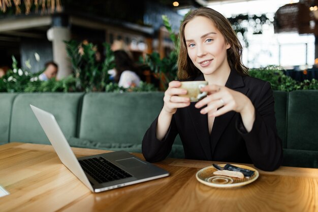 カフェテリアでの若い美しさの女性はノートパソコンと電話で話し、コーヒーを飲みます。