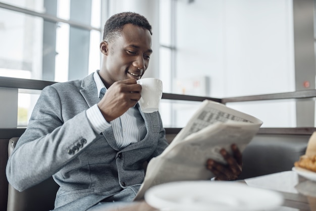 オフィスカフェで昼食をとっている新聞を持つ若い黒人実業家。成功したビジネスパーソンはフードコートでコーヒーを飲み、正装で黒人男性 Premium写真