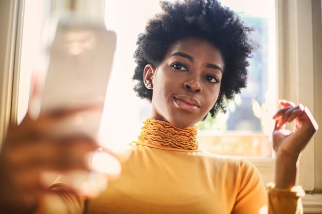 Young black female with an orange sweater taking a selfie on a roof in front of a window