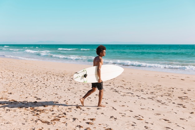 Молодой черный мужчина идет с доской для серфинга в море