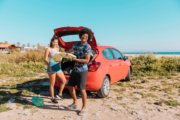 Young black man holding surfboard near car by seashore