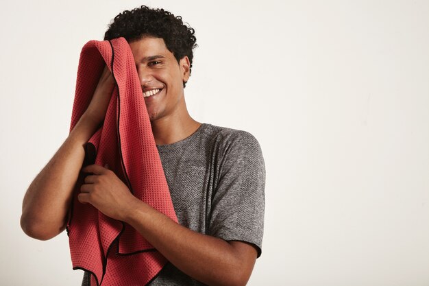 Young black sportsman laughs and wipes his face with a red towel, right half of the face open on white