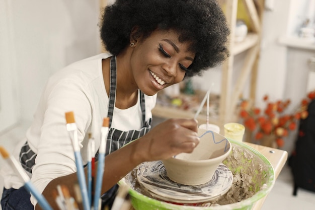 Free photo young black woman painting clay pot at art studio craftswoman painting ceramic bowl on workbench girl wearing a blue apron