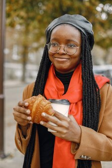 긴 loc 헤어스타일을 가진 젊은 흑인 여성이 커피 한 잔과 크루아상을 들고 밖에 서서 갈색 코트 오렌지 스카프와 검은 모자를 쓴 여성