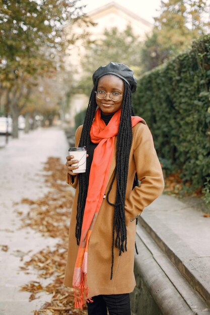 긴 loc 헤어스타일을 한 젊은 흑인 여성이 테이크 아웃 커피 한 잔을 들고 밖에 서 있습니다. 갈색 코트, 주황색 스카프, 검은 모자를 쓴 여성