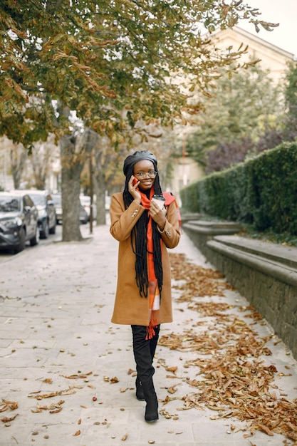 Young black woman with a long locs hairstyles standing outside with a cup of take away coffee. Woman wearing brown coat, orange scarf and black hat