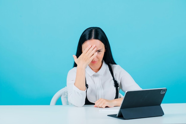 Young blogger girl is looking at tablet by holding hand on forehead on blue background