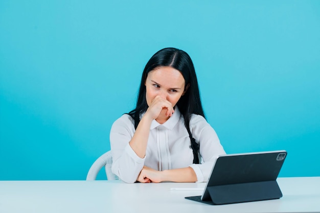 Young blogger girl is posing to tablet camera by holding hand on nose on blue background