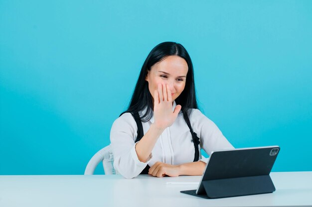 Young blogger girl is showing hi gesture to tablet camera on blue background