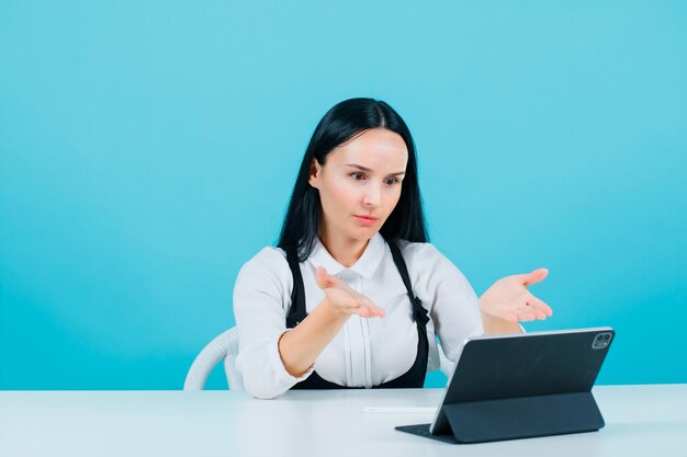 Young blogger girl is talking on video call with table by extending hands to camera on blue background