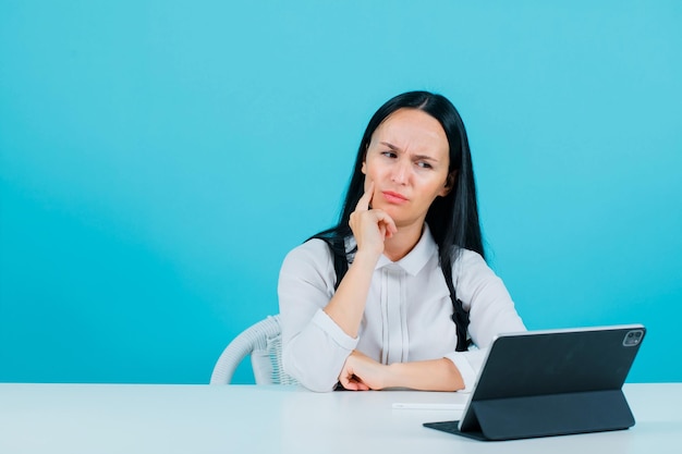 Young blogger girl is thinking by holding hand on cheek and sitting in front of tablet camera on blue background