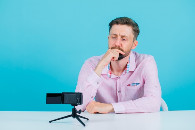 Young blogger man is looking at his mini camera by holding ahnd on mouth on blue background