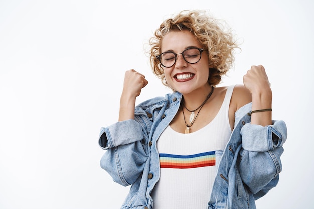 young blond girl in rainbow top and denim jacket clenching raised fists in celebration and triumph smiling, close eyes from happiness.