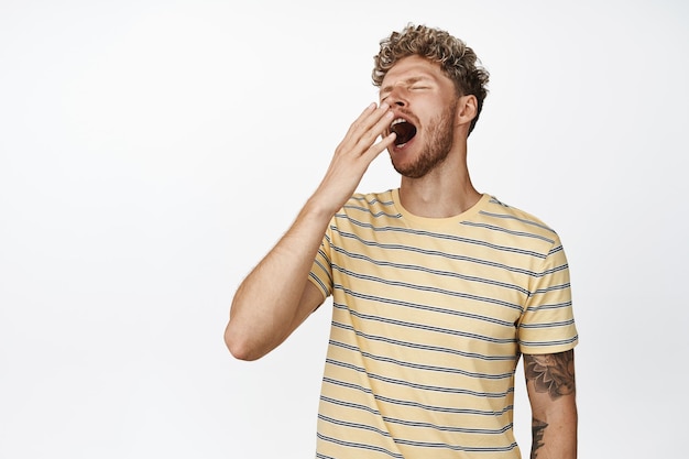 Young blond handsome man yawning from fatigue standing sleepy cover yawn with hand near opened mouth standing over white background waking up early