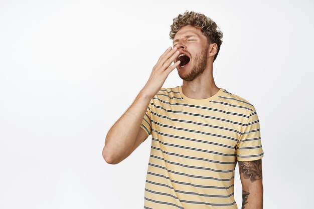 Young blond handsome man yawning from fatigue standing sleepy cover yawn with hand near opened mouth standing over white background waking up early