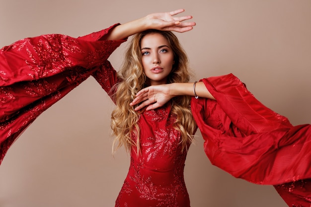 young blond lovely woman in luxurious red dress with wide sleeves. Expressive pose.