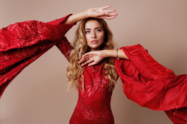 young blond lovely woman in luxurious red dress with wide sleeves. Expressive pose.
