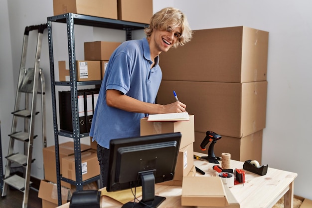 Young blond man ecommerce business worker using laptop writing on notebook at office