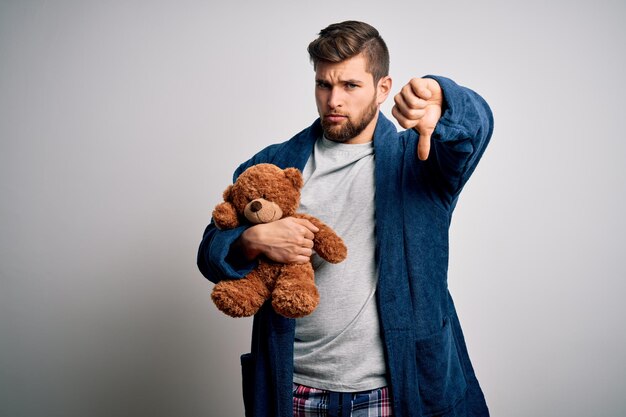 Young blond man with beard and blue eyes wearing pajama holding teddy bear with angry face negative sign showing dislike with thumbs down rejection concept