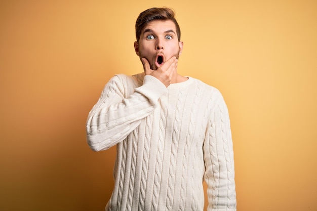 Young blond man with beard and blue eyes wearing white sweater over yellow background Looking fascinated with disbelief surprise and amazed expression with hands on chin