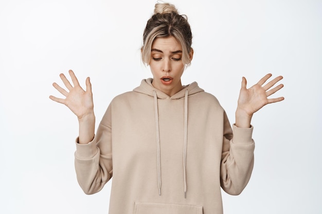 Young blond silly girl drop something raise hands and look down concerned standing against white background