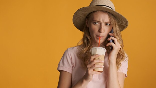 Young blond woman in hat drinking coffee while have serious phone conversation over colorful background. Copy space