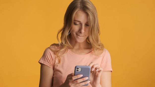 노란색 배경에 격리된 스마트폰으로 메시지를 열심히 읽는 젊은 금발 여성. 소셜 미디어 서핑