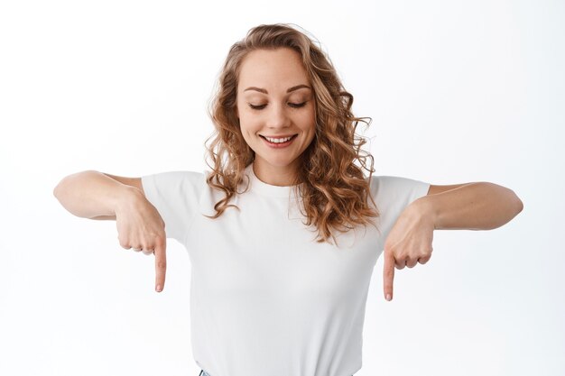 Young blond woman looking down and pointing copyspace, showing advertisement, standing over white wall