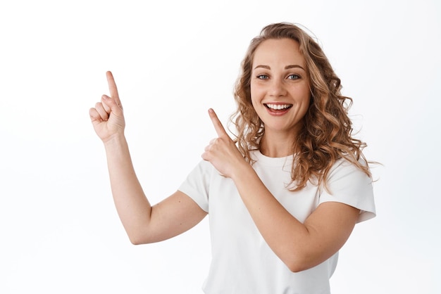 Young blond woman smiling cheerful pointing at upper left corner copyspace showing advertisement standing over white background