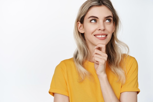 Young blond woman smiling thinking and looking aside with satisfied face has an idea standing in yellow tshirt against white background