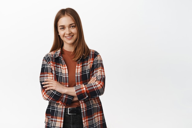 Young blond wonan in plaid shirt, cross arms on chest and smiling with confidence standing over white wall.