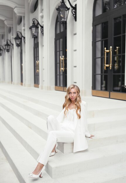 Young blonde curly haired woman in Classic style white suit sitting posing on stairs Beautiful caucasian bride model girl in wedding fashionable clothes Wedding style