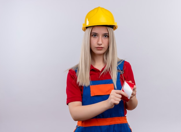 Free photo young blonde engineer builder girl in uniform holding scotch tape looking  on isolated white wall with copy space