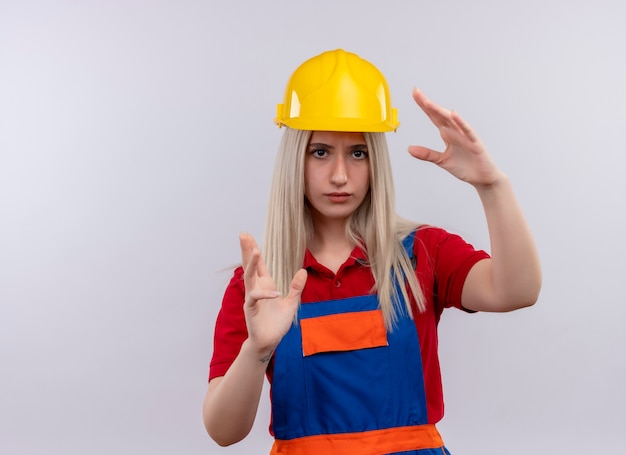 Giovane ragazza bionda del costruttore dell'ingegnere in uniforme che mostra la forma di qualcosa che osserva sulla parete bianca isolata con lo spazio della copia