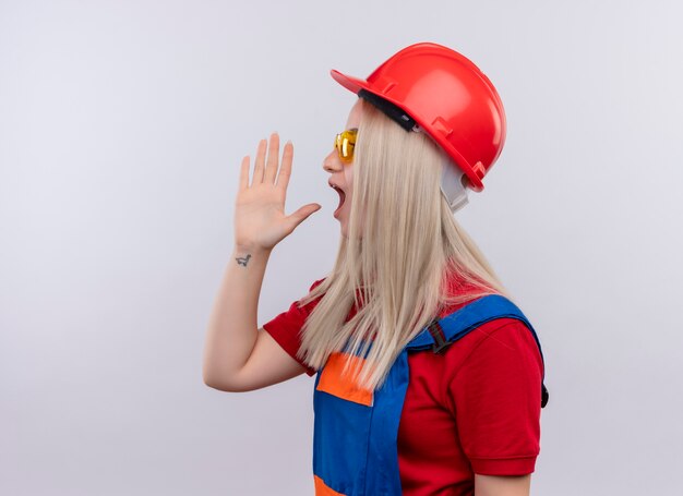 Молодая блондинка инженер-строитель девушка в униформе в защитных очках кричит на кого-то с рукой возле рта, стоящего в профиле на изолированной белой стене с копией пространства