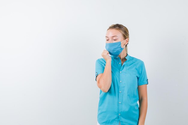 Young blonde female in casual clothes, mask holding hand near mouth while looking down