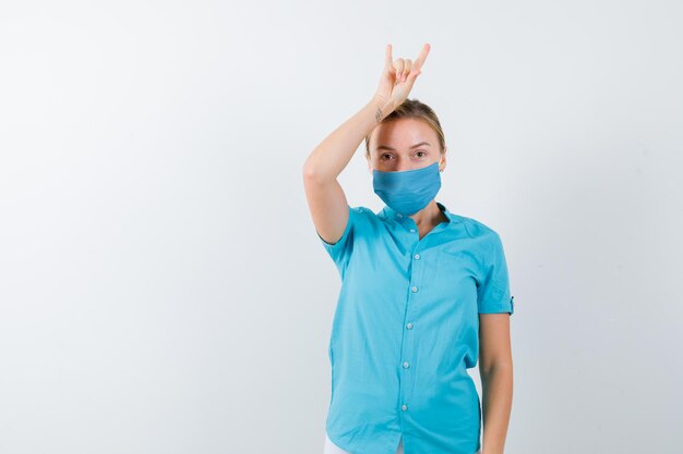 Young blonde female showing rock gesture in casual clothes, mask and looking merry