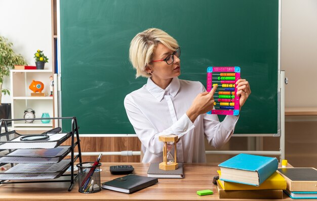 young blonde female teacher wearing glasses sitting at desk with school supplies in classroom showing abacus looking at it pointing finger on it