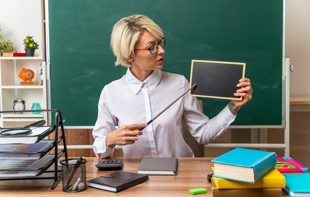 Foto gratuita giovane insegnante di sesso femminile bionda con gli occhiali seduto alla scrivania con gli strumenti della scuola in aula che mostra la mini lavagna guardandola puntata con il bastone del puntatore