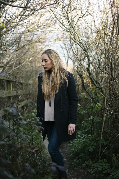 Young blonde female with a black coat standing on a pathway surrounded by leafless trees